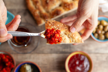 Healthy Turkish breakfast, with sliced tomato, cucumber, and white feta cheese, small bowls of strawberry jam, olive, honey, pepper paste. Woman is putting pepper paste onto slice of pide.