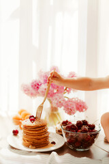 child's hand pricks a stack of pancakes with fresh cherries on top with a gold fork. There is bowl of cherries and apricots, pink hyacinth flowers on the white background. Vertical photo, Copy space.