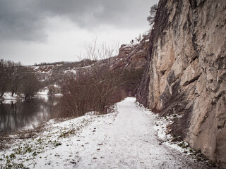 Fototapeta na wymiar White footpath full of snow and without people. On one sight rocky hill and on the other slowly flowing river.
