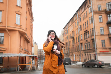 Obraz premium Happy young woman with glass of coffee in her hands, talking on phone on walk.