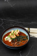 Asian miso soup with tofu, tuna, shiitake mushrooms, seaweed, sesame seeds and spring onion. Dish isolated in a blue bowl, close-up on a black marble background. Asian cuisine. Copy space.