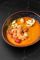 Asian soup Tom Yam with shrimps, mussels, mushrooms, tomatoes and spring onion. Dish isolated in a blue bowl, close-up on a black marble background. Asian cuisine.