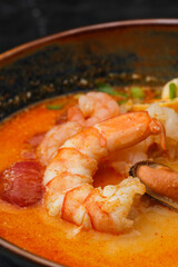 Asian soup Tom Yam with shrimps, mussels, mushrooms, tomatoes and spring onion. Dish isolated in a blue bowl, close-up on a black marble background. Asian cuisine.