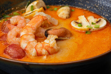 Asian soup Tom Yam with shrimps, mussels, mushrooms, tomatoes and spring onion. Dish isolated in a blue bowl, close-up on a black marble background. Asian cuisine.