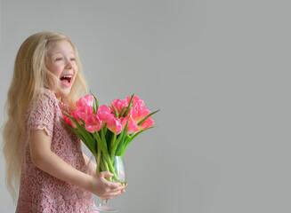 Laughing cute little blonde girl holding a vase of pink tulips on the grey background