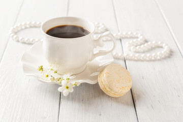 One beige macaroon and cup of hot coffee against pearl beads on a white wood table. Sweet breakfast with delicious french dessert. Valentine's day greeting card. High key image. Selective focus.