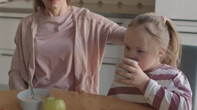 Medium Shot Of Unrecognizable Woman Sitting At Kitchen Table And Patting On Head Her Lovely Little Daughter With Down Syndrome While She Drinking Juice During Breakfast