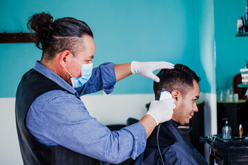 Fototapeta premium hispanic man stylist with facemask cutting hair to a client in a barber shop in Mexico city in coronavirus pandemic
