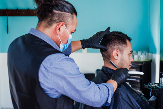 hispanic man stylist with facemask cutting hair to a client in a barber shop in Mexico city in coronavirus pandemic