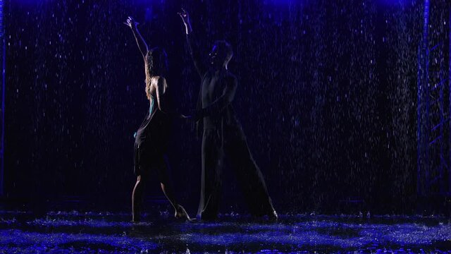 Ballroom dancers dancing passionate Latin American bachata dance in pouring rain. Silhouettes of a young couple in wet clothes on black studio background with water drops and blue light. Slow motion.