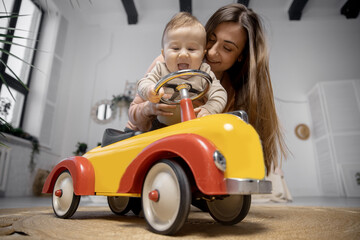 Concept Young baby driver. Mother plays with her little son and big toy car, boy holds steering wheel and laughs