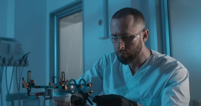 A model of an upper jaw in axiograph face-bow is standing on the table in a dark laboratory; a male dentistry worker is sitting next to it, checking and exploring the device.