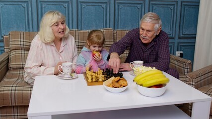 Senior grandparents and child granddaughter spending time home together, sitting, playing chess game