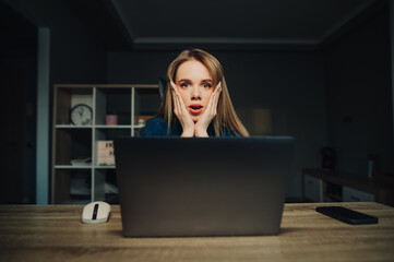 Portrait of a shocked female remote worker sitting with a laptop at home at the table and looking at the screen with an emotional face. Surprised freelancer woman working at home