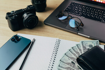 top view of office desk workplace with camera, notebook, glasses, laptop, smartphone and dollars banknotes on wooden table background.