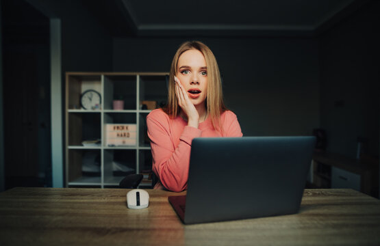 Portrait of shocked female online worker sitting at desk at home with laptop and looking at camera with surprised face.