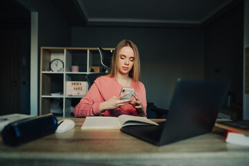 Cute female student sitting at home at desk with laptop and books and uses smartphone with serious face in pause between studies.