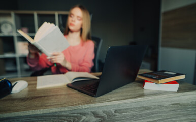 Background. Photo in defocus. A female student studies remotely at home, holds a book in her hands and uses a laptop.