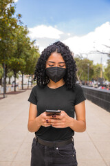 Young woman with curly hair uses her phone, she has a black mask on her face