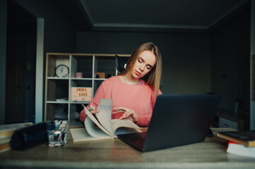 Obraz premium Female student in a pink sweater studies remotely at home, looks in a notebook with a serious face and uses a laptop. Home learning concept.