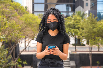 Young woman with curly hair uses her phone, she is sitting and has a black mask