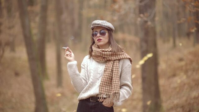 Autumn fashion vogue. Stylish hipster girl wearing modern dress in urban park and smoking cigarette. Female model.
