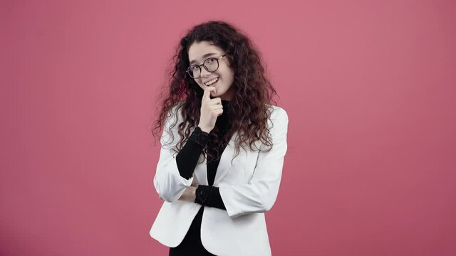 The student with curly hair, the eminence looks into the room with her hands to her lips smiling ironically. Young hipster in white jacket and black shirt, with glasses posing isolated on pink