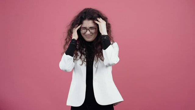 The young woman with curly hair has migraines, massaging with her hands. Young hipster in white jacket and black shirt, with glasses posing isolated on pink background in the studio. The concept of