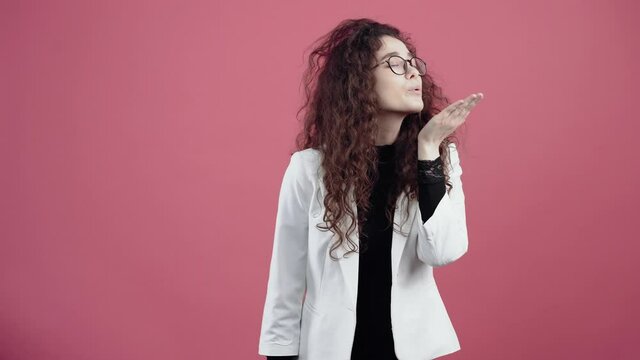 The young woman in love, shares kisses blowing in the palm left and right. Young hipster in white jacket and black shirt, with glasses posing isolated on pink background in the studio. The concept of