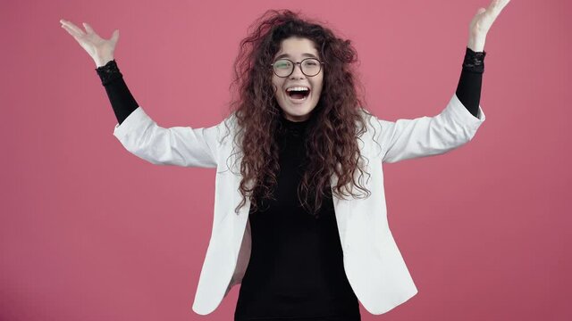 Shocked young woman with curly hair, she smiles happily putting her hands in her hair. Young hipster in white jacket and black shirt, with glasses posing isolated on pink background in the studio. The