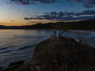 Fototapeta premium Beautiful and peaceful aerial view of a woman practicing yoga and meditation on a rock over the sunset in a beach 