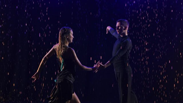 Passionate bachata dance among the raindrops performed by a young sensual couple of dancers. The partners move in a dance on a black studio background among blue neon lights. Close up. Slow motion.