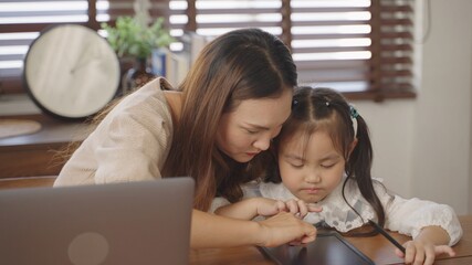 Young asian businesswoman mother using the laptop for working online from home with cute little girl drawing on tablet in the morning during quarantine of global coronavirus epidemic.