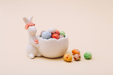 Fototapeta na wymiar Easter chocolate mini candies in the ceramic stand with bunny figurine on a beige background