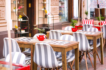Fototapeta na wymiar red teapot flowerpot with flower on wooden table terrace cafe with striped soft seats near facade old building, nobody.
