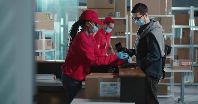 On pandemic post office sevices multi-ethnic people staff in medical masks giving order for caucasian customer client receiving parcel delivery during quarantine.