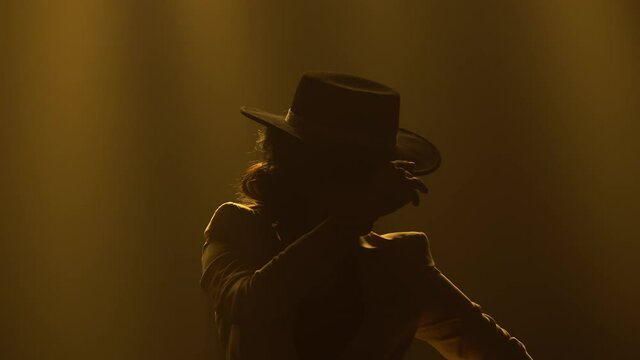 Stylish sexy woman dancing flamenco elements in a dark smoky studio in the rays of yellow light. A silhouette of a dancer in a trouser suit and a hat moves passionately in the dance. Close up.