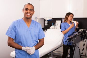 Positive male doctor cosmetologist meeting client in office while female assistant preparing equipment for procedures in background