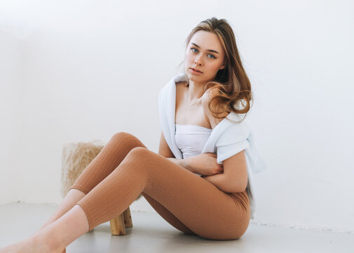 Young woman in white hoodie on shoulders beautiful fit girl in sport wear sitting on floor in the white studio