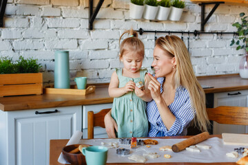 Funny little baby helper playing with dough on his hands learning to knead helps adult mom in the kitchen, happy cute baby daughter and parent mom have fun cooking cookies.