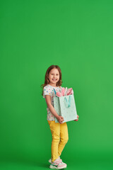 cute little girl in stylish clothes, wearing a T-shirt with colorful polka dots, stands with a bag with bunny ears on a green background on Easter day. Full-length. Copy space.