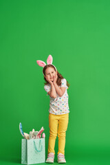 Cute little girl in stylish clothes, wearing Easter bunny ears, goes with a bag with bunny ears on a green background on Easter day. Full-length. Copy space.