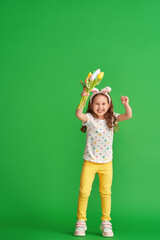 charming happy girl with Easter bunny ears jumps on a green background in full growth. a child smiles and holds a bouquet of fabric tulip flowers on Easter Day. The joy of the arrival of spring.