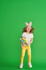 charming happy girl with Easter bunny ears jumps on a green background in full growth. a child smiles and holds a bouquet of fabric tulip flowers on Easter Day. The joy of the arrival of spring.