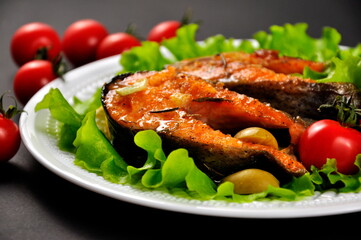 Serving. Baked trout on a white plate, with lemon and cherry tomatoes. Black background.