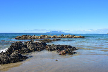 Fototapeta na wymiar Flat formation of dark rocks exposed on sandy beach during low tide with island far in background.