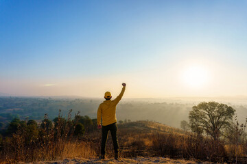 Back view of a young male wearing a yellow hoodie and cap with his fist up in a field at sunset
