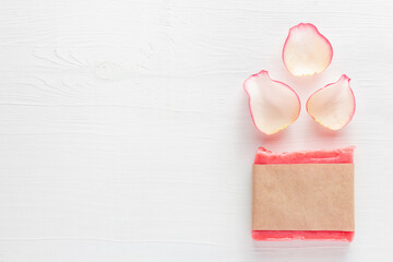 Pink handmade floral soap bar in blank paper label package next to rose petals on white table background. Skin care cosmetic product mockup. Zero waste spa concept.