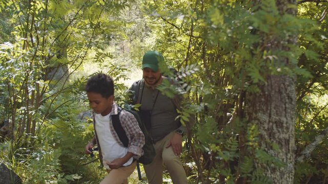 Smiling Caucasian father and little mixed race son walking with backpacks through forest while hiking together on summer day