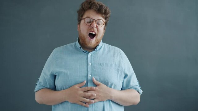 Portrait of funny bored guy yawning looking at camera standing against gray background with unhappy face. Millennials and human emotions concept.